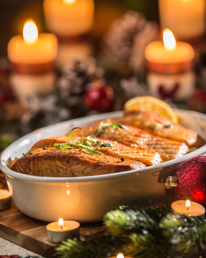 Filés de salmão assados em travessa, decorados com alecrim e laranja, entre velas de Natal