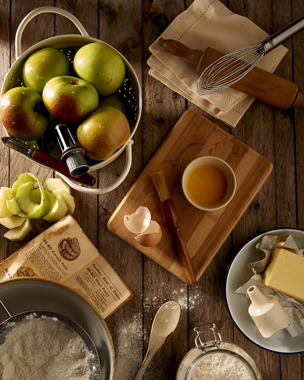 Ingredientes separados sobre a bancada de madeira para o preparo de uma torta: maçãs, farinha, manteiga, ovos e utensílios